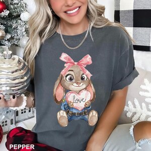 May include: Dark gray t-shirt featuring a cartoon bunny holding a heart-shaped sign with the word "Love" on it. The bunny has a pink bow and is wearing a blue uniform. The shirt is a comfortable, casual style.