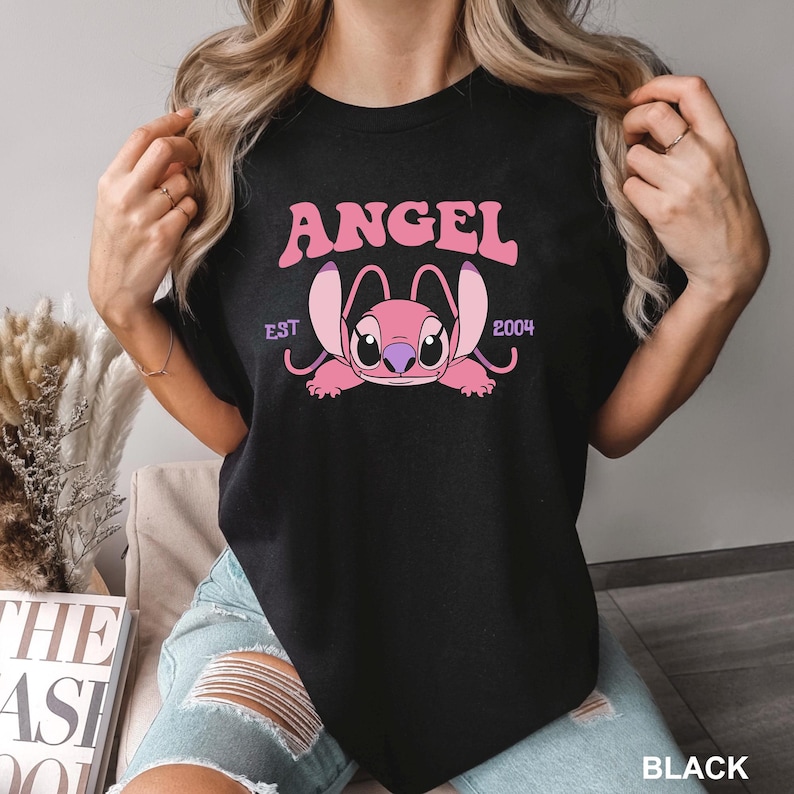 K&ouml;nnte beinhalten: Schwarzes T-Shirt mit einem rosa Aufdruck der Figur Angel aus Lilo & Stitch. Das Wort "ANGEL" steht &uuml;ber der Figur, darunter "EST 2004". Das T-Shirt hat einen Rundhalsausschnitt.