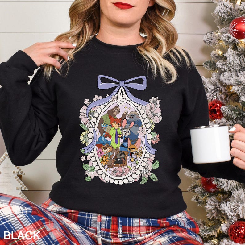May include: Black crewneck sweatshirt featuring a floral oval design with cartoon characters. The design includes a light purple bow at the top and is surrounded by flowers in shades of pink, white, and green. The word "BLACK" is printed at the bottom.