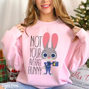 Op de afbeelding: Lichtroze sweatshirt met de tekst "NOT YOUR AVERAGE BUNNY" en een cartoonkonijn. Het konijn draagt een politie-uniform en houdt een aktetas vast. De sweatshirt heeft een ronde hals.