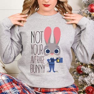 Op de afbeelding: Sport Grey sweatshirt met de tekst "NOT YOUR AVERAGE BUNNY" en een cartoon konijn. Het konijn heeft grijs haar, roze oren en draagt een blauw uniform. De sweatshirt is lichtgrijs.