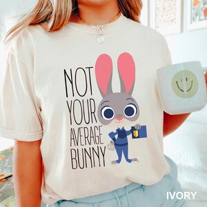 May include: Cream-coloured t-shirt with the text "NOT YOUR AVERAGE BUNNY" and a cartoon bunny in a police uniform. The bunny has pink ears and is holding a badge and notepad. A white mug with a smiley face is also visible.
