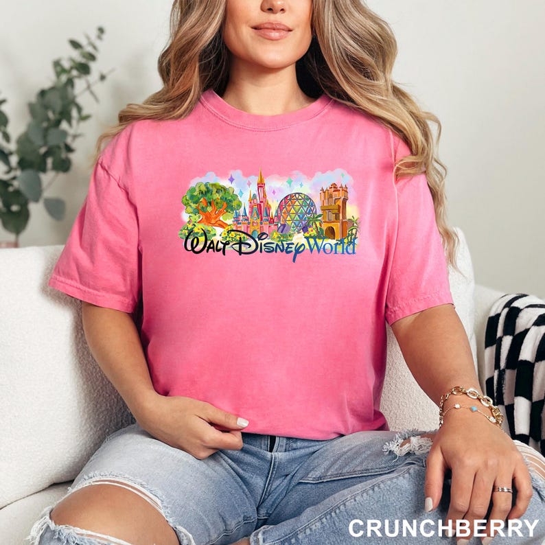 May include: A bright pink short-sleeved t-shirt featuring a colorful graphic print of Walt Disney World landmarks, including a castle, a geodesic dome, and a tree, with the text Walt Disney World printed in a stylized blue script font across the front.