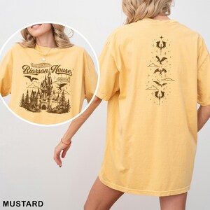 May include: Mustard-colored t-shirt featuring a detailed graphic of a castle with the text "Riorrson House" and "Aretia". The back of the shirt displays a series of dragon illustrations. The shirt is a relaxed fit.