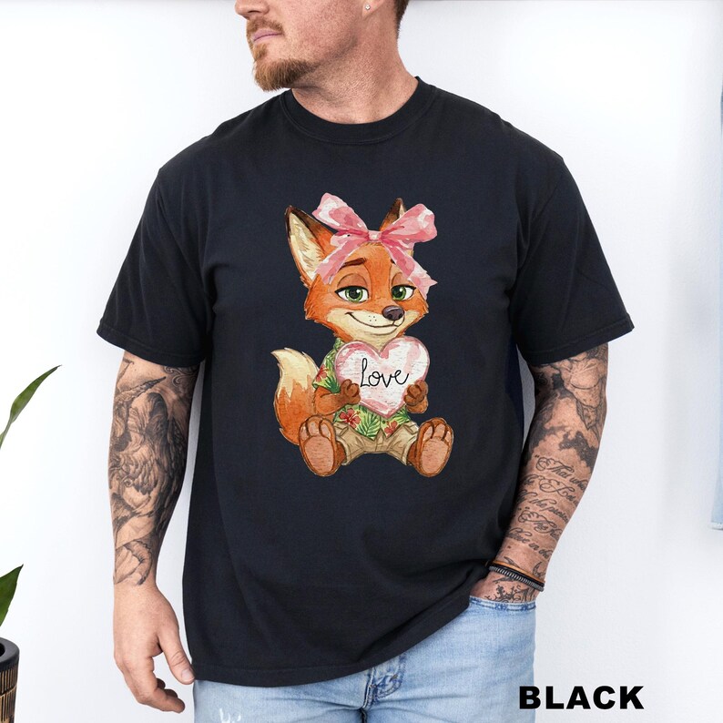 May include: Black t-shirt featuring a cartoon fox with a pink bow, holding a heart-shaped sign that says "Love." The fox is wearing a Hawaiian shirt and khaki shorts. The word "BLACK" is printed at the bottom.