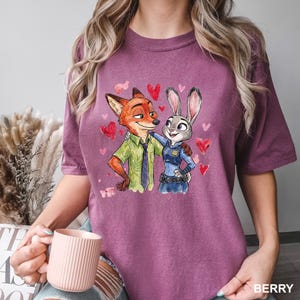 Może przedstawiać: Koszulka w kolorze jagodowym z akwarelową ilustracją Nicka Wilde'a i Judy Hopps z Zootopii, otoczonych czerwonymi sercami. Nick nosi zieloną koszulę, a Judy mundur policjantki. Słowo "BERRY" jest wydrukowane na dole.