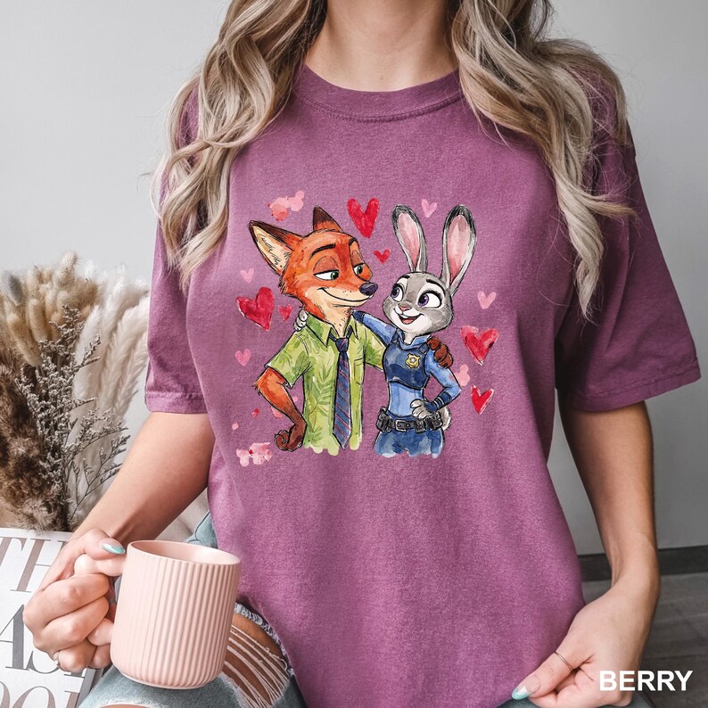 Może przedstawiać: Koszulka w kolorze jagodowym z akwarelową ilustracją Nicka Wilde'a i Judy Hopps z Zootopii, otoczonych czerwonymi sercami. Nick nosi zieloną koszulę, a Judy mundur policjantki. Słowo "BERRY" jest wydrukowane na dole.