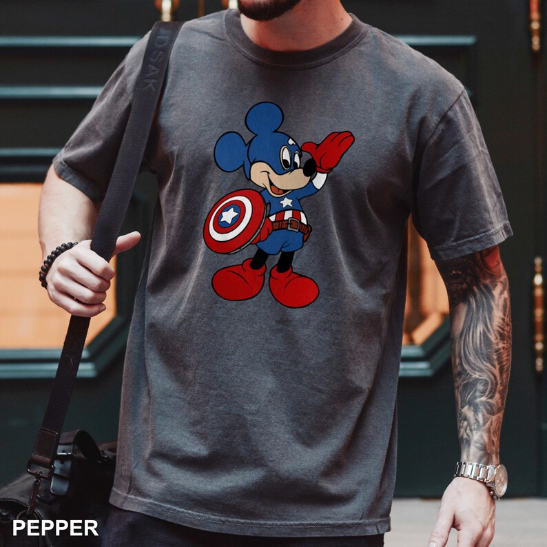 May include: Dark gray t-shirt featuring a cartoon Mickey Mouse dressed as Captain America. Mickey is wearing a blue costume, holding a shield, and saluting. The shirt has a distressed look, with the word "PEPPER" printed at the bottom.