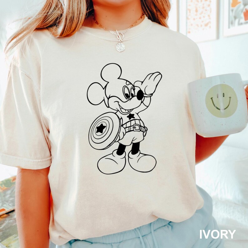 May include: Ivory-colored t-shirt featuring a black and white graphic of Mickey Mouse as Captain America, holding a shield with a star. The shirt has short sleeves. A white mug with a smiley face is also visible.