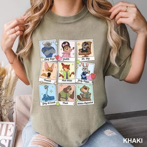Op de afbeelding: Een kaki t-shirt met ronde hals voorzien van een raster van negen illustraties in polaroidstijl van animatiefiguren. Elk kader bevat een naam: Chief Bogo, Judith, Mr. Big, Finnick, Nick Wilde, Judy Hopps, Gary de'snack, Flash en Nibbles Maplestick.