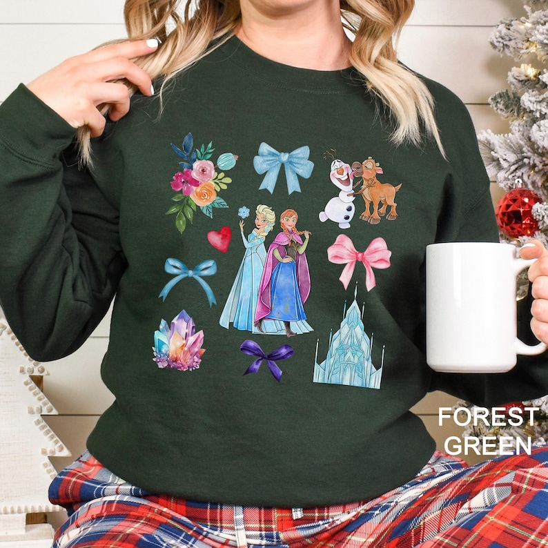 以下が含まれることがあります： 人気の長編アニメ映画のキャラクターと要素のカラフルなグラフィックが特徴のフォレストグリーンのスウェットシャツ。デザインには、2人のプリンセス、雪だるま、トナカイ、リボン、花、お城のイラストが含まれています。「FOREST GREEN」の文字が右下にプリントされています。