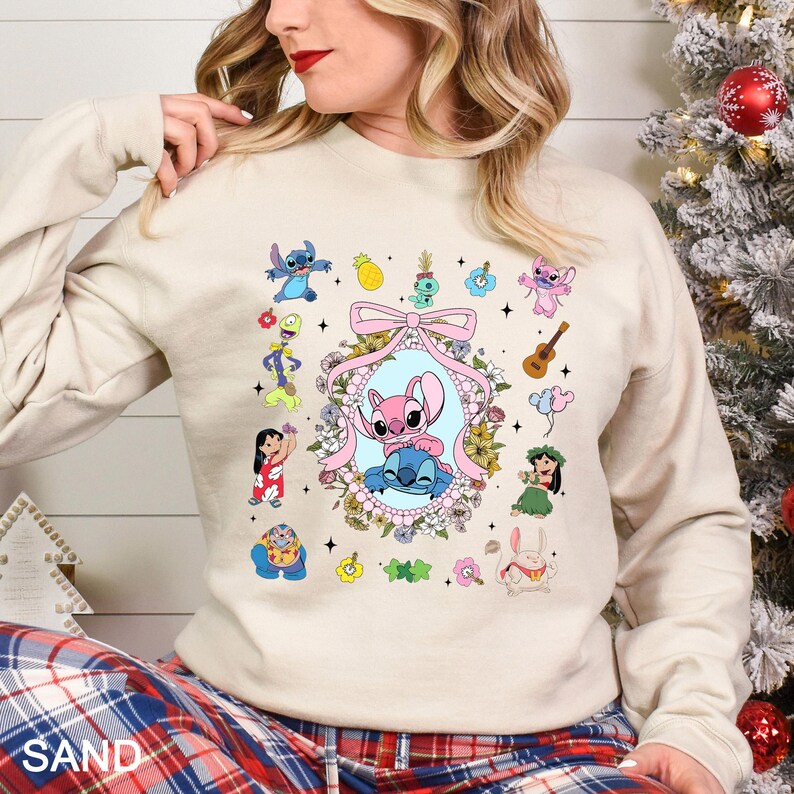May include: A sand-colored sweatshirt featuring a colorful graphic of Stitch and Angel, surrounded by other characters and floral elements. The design includes a pink bow and the word "SAND" at the bottom.