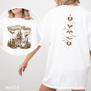 May include: White t-shirt with a brown graphic on the front featuring a castle, dragons, and text that reads "Riorson House". The back of the shirt has a vertical design of dragons and decorative elements. The shirt is a casual style.