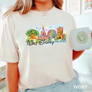 May include: An ivory short-sleeve t-shirt featuring a colorful graphic of Walt Disney World landmarks, including a tree, a castle, a geodesic dome, and a tower, with the text Walt Disney World in black script lettering across the center.
