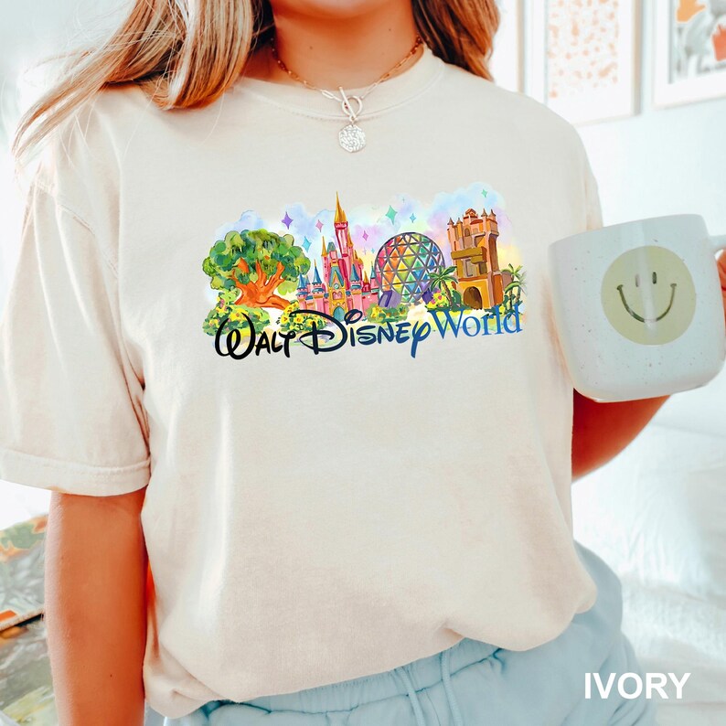 May include: An ivory short-sleeve t-shirt featuring a colorful graphic of Walt Disney World landmarks, including a tree, a castle, a geodesic dome, and a tower, with the text Walt Disney World in black script lettering across the center.