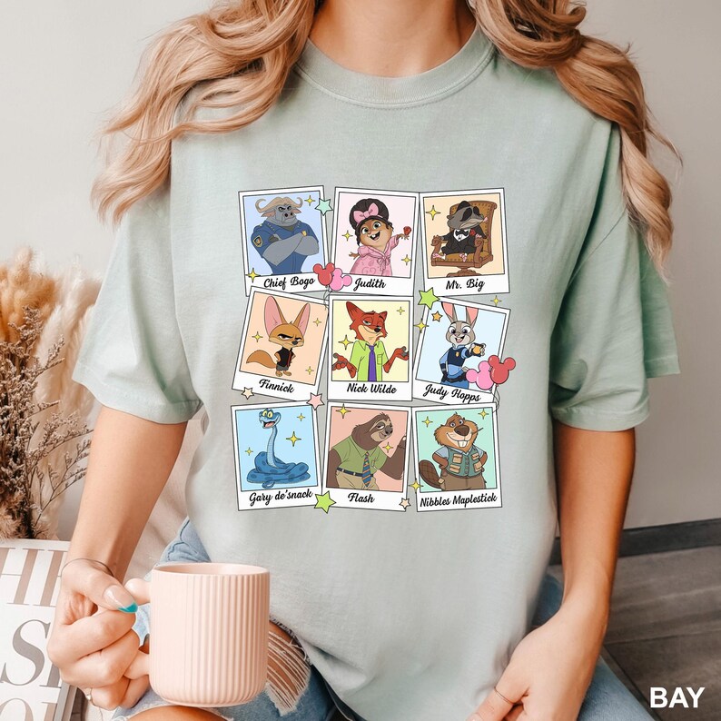 Op de afbeelding: Een licht saliegroen t-shirt met een raster van negen polaroid-achtige kaders. Elk kader bevat een stripfiguur met de naam eronder gedrukt: Chief Bogo, Judith, Mr. Big, Finnick, Nick Wilde, Judy Hopps, Gary de'snack, Flash en Nibbles Maplestick.