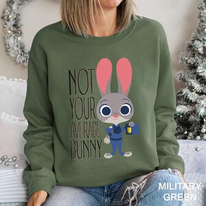 Op de afbeelding: Militairgroen sweatshirt met een cartoon konijn ontwerp. Het konijn heeft roze oren en draagt een blauw uniform, met een blauwe tas. De tekst luidt "NOT YOUR AVERAGE BUNNY".