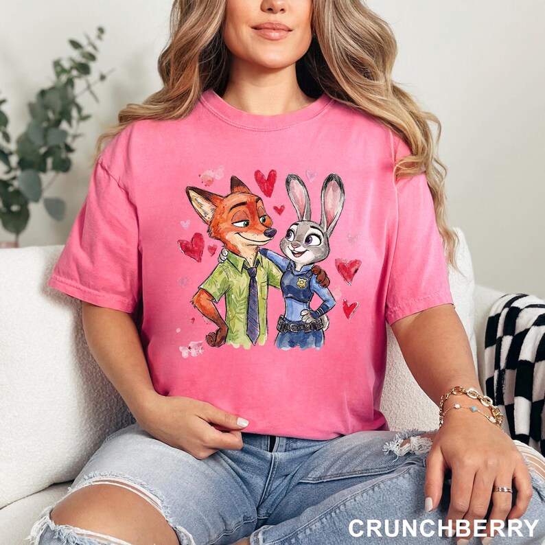 Może przedstawiać: R&oacute;żowa koszulka z akwarelową ilustracją Nicka Wilde'a i Judy Hopps z Zootopii, otoczona czerwonymi sercami. Nick ma na sobie zieloną koszulę i krawat, Judy mundur policjantki. Słowo "CRUNCHBERRY" znajduje się na dole.