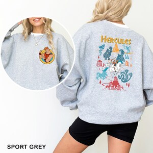 K&ouml;nnte beinhalten: Sportgraues Sweatshirt mit einer Cartoon-Illustration von Herkules auf der Vorder- und R&uuml;ckseite. Die Vorderseite zeigt Herkules beim Posen, w&auml;hrend die R&uuml;ckseite ein farbenfrohes Design mit dem Wort "Hercules" und Szenen aus dem Film zeigt.