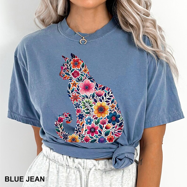May include: Blue jean colored t-shirt featuring a floral cat design. The cat silhouette is filled with colorful flowers in shades of pink, orange, yellow, and blue. The text "BLUE JEAN" is printed at the bottom of the shirt.