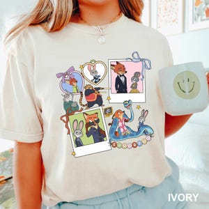 May include: Ivory t-shirt with a colourful graphic of "Zootopia" characters in polaroid frames. The design includes animal characters, stars, and the word "Zootopia". A white mug with a smiley face is held in the image.