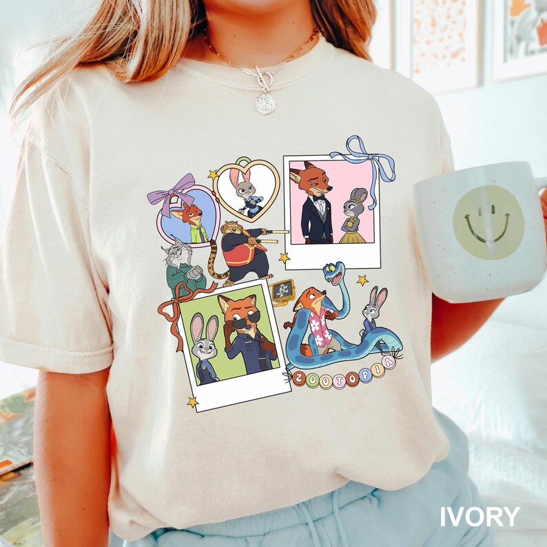 May include: Ivory t-shirt with a colourful graphic of "Zootopia" characters in polaroid frames. The design includes animal characters, stars, and the word "Zootopia". A white mug with a smiley face is held in the image.