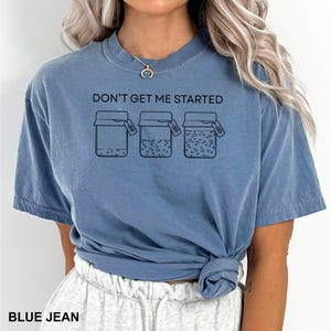 May include: Blue jean colored t-shirt with the text "DON'T GET ME STARTED" above three jars with varying contents. The bottom of the shirt has the text "BLUE JEAN". The shirt is a short-sleeved, crew neck style.