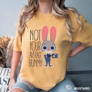 May include: Mustard-coloured t-shirt with a cartoon bunny in a police uniform and the text "NOT YOUR AVERAGE BUNNY". The bunny has pink ears and a briefcase. The shirt is a solid colour.