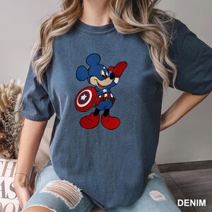 May include: Denim blue t-shirt featuring a cartoon Mickey Mouse dressed as Captain America. Mickey is wearing a blue costume, holding a red and white shield, and saluting. The word "DENIM" is in the bottom right corner.