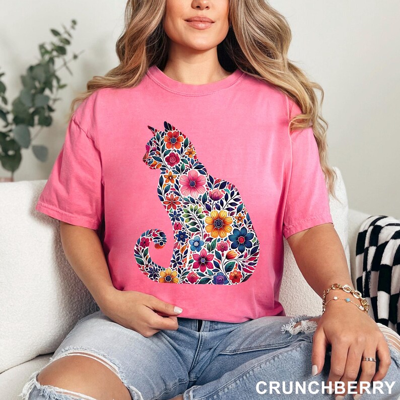 May include: A pink t-shirt featuring a floral cat design. The cat silhouette is filled with colorful flowers in shades of pink, blue, yellow, and orange. The text "CRUNCHBERRY" is visible at the bottom right.