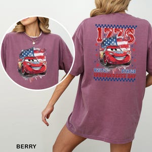 May include: A berry-coloured t-shirt featuring a graphic of a red race car in front of an American flag. The back design includes the text 1776 and UNITED STATES INDEPENDENCE DAY. The front chest area displays a smaller version of the same graphic.
