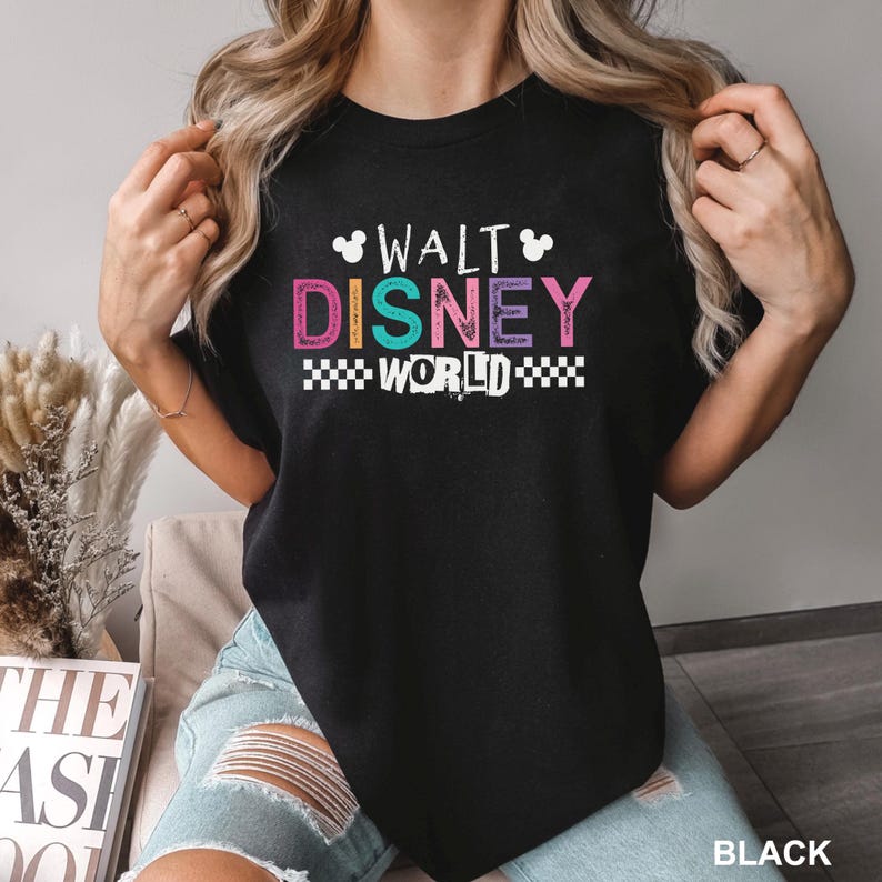 May include: A black short-sleeved t-shirt featuring the text WALT DISNEY WORLD in a multi-colored, distressed font. The design includes small white Mickey Mouse head silhouettes above the text and a black and white checkered pattern below.