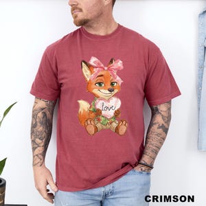 May include: Crimson-colored t-shirt featuring a cartoon fox with a pink bow, holding a heart-shaped sign that says "love". The fox is wearing a green shirt and khaki shorts. The shirt is a comfortable, casual style.
