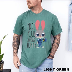 May include: Light green t-shirt featuring the text "NOT YOUR AVERAGE BUNNY" and a cartoon bunny character. The bunny has grey fur, pink ears, and is wearing a blue uniform and holding a briefcase. The shirt is a solid colour.