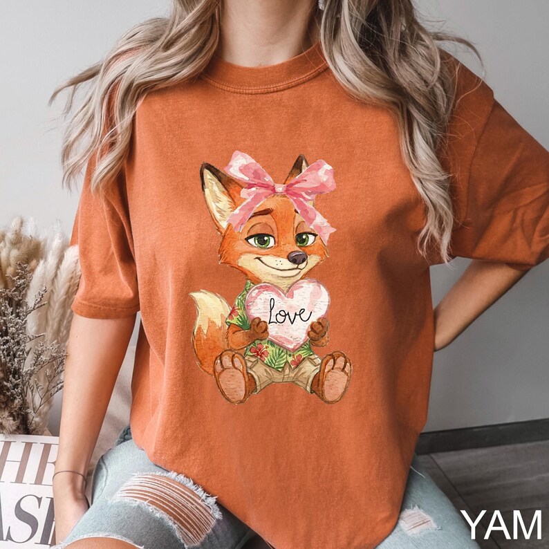 May include: Burnt orange t-shirt featuring a cartoon fox holding a heart-shaped sign with the word "Love". The fox has a pink bow and is wearing a Hawaiian shirt. The shirt has short sleeves.