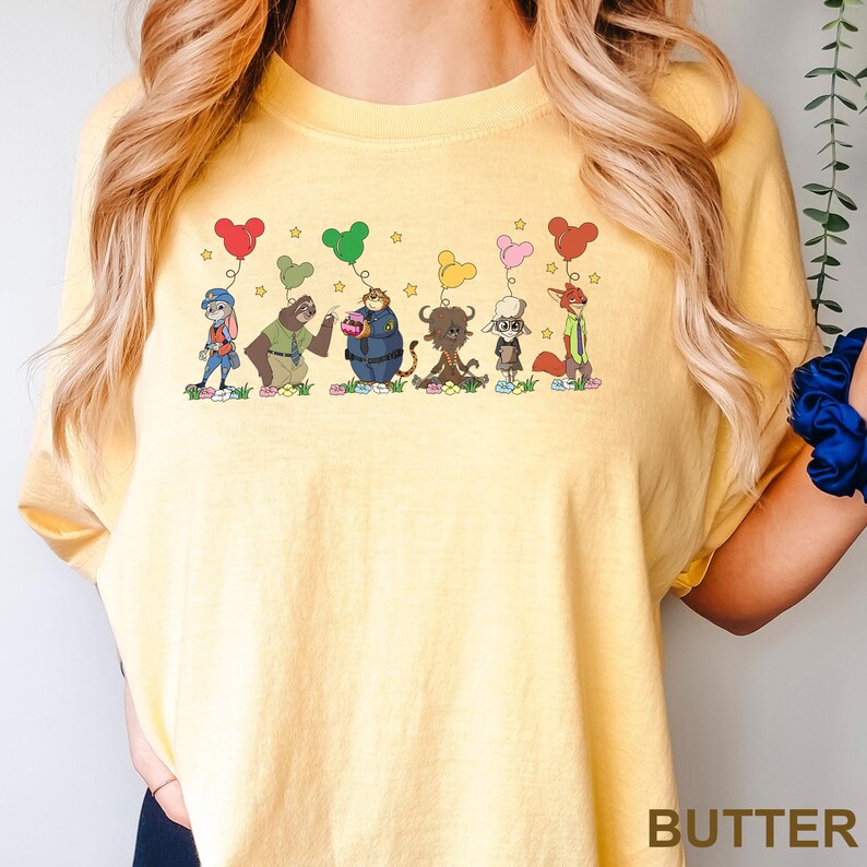 May include: Yellow t-shirt with a cartoon graphic of various animal characters holding balloons. The word "BUTTER" is printed in the bottom right corner. The graphic features a sloth, a police officer, and a fox.