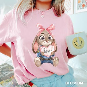 May include: A light pink t-shirt featuring a cartoon bunny holding a heart-shaped sign with the word "Love". The bunny has a pink bow and is wearing a police uniform. A white mug with a yellow smiley face is held in the hand.