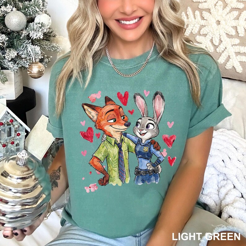 Może przedstawiać: Jasnozielona koszulka z kresk&oacute;wkową ilustracją Nicka Wilde'a i Judy Hopps z Zootopii, otoczona czerwonymi sercami. Tekst "LIGHT GREEN" znajduje się na dole. Postacie się obejmują, Nick ma na sobie zieloną koszulę, a Judy mundur policjantki.