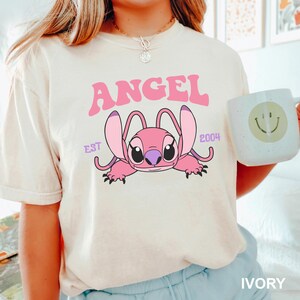 K&ouml;nnte beinhalten: Elfenbeinfarbenes T-Shirt mit einer rosa Cartoonfigur und dem Wort "ANGEL". Die Figur hat gro&szlig;e Ohren und liegt. Der Text "EST 2004" steht ebenfalls auf dem Shirt. Eine wei&szlig;e Tasse mit einem Smiley wird im Bild gehalten.