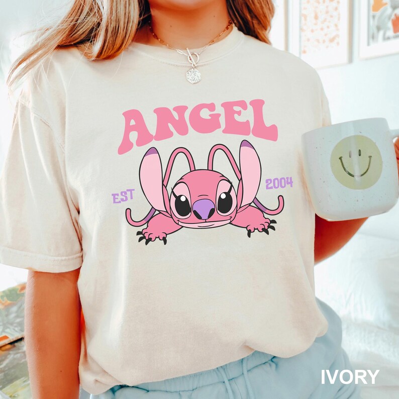 K&ouml;nnte beinhalten: Elfenbeinfarbenes T-Shirt mit einer rosa Cartoonfigur und dem Wort "ANGEL". Die Figur hat gro&szlig;e Ohren und liegt. Der Text "EST 2004" steht ebenfalls auf dem Shirt. Eine wei&szlig;e Tasse mit einem Smiley wird im Bild gehalten.
