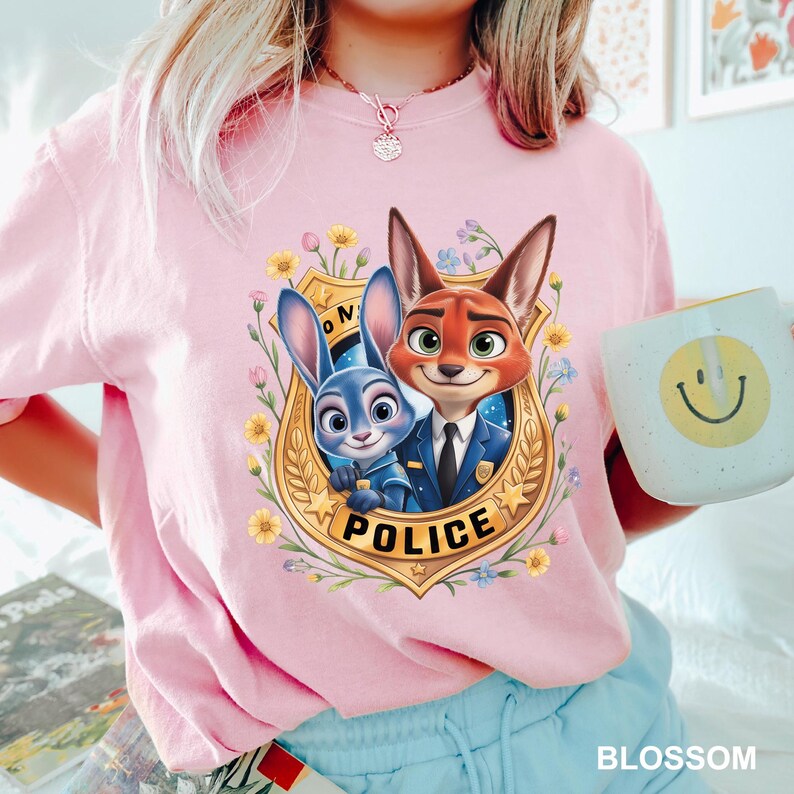 Peut inclure: Un t-shirt rose clair avec un dessin anim&eacute; de deux personnages de Zootopie &agrave; l'int&eacute;rieur d'un badge de police, entour&eacute; de fleurs. Le mot "POLICE" est imprim&eacute; sur le badge. Une tasse blanche avec un smiley jaune est tenue dans la main.