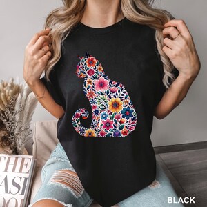 May include: Black t-shirt featuring a floral cat silhouette. The cat design is filled with colorful flowers in shades of pink, yellow, blue, and orange. The word "BLACK" is printed at the bottom right corner of the shirt.