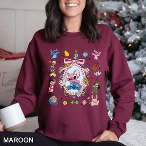 May include: Maroon sweatshirt featuring a colorful graphic of cartoon characters. The design includes a central oval with two characters, surrounded by flowers and a pink bow. Additional character illustrations are scattered around the main design.