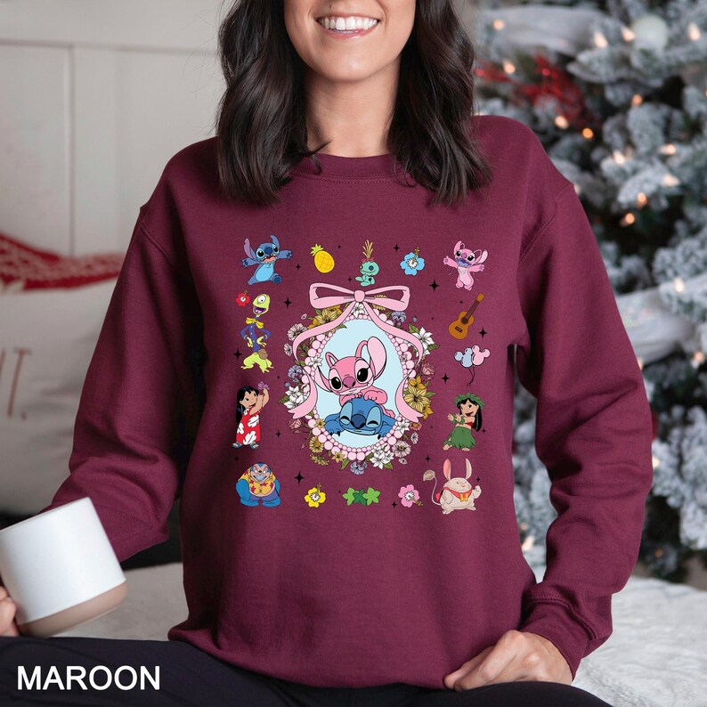 May include: Maroon sweatshirt featuring a colorful graphic of cartoon characters. The design includes a central oval with two characters, surrounded by flowers and a pink bow. Additional character illustrations are scattered around the main design.