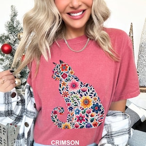 May include: Crimson-colored t-shirt featuring a floral cat silhouette. The cat design is filled with colorful flowers in shades of pink, orange, blue, and yellow. The word "CRIMSON" is printed at the bottom of the shirt.