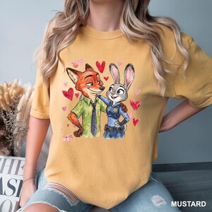 Może przedstawiać: Musztardowa koszulka z akwarelową ilustracją Nicka Wilde'a i Judy Hopps z Zootopii, otoczona r&oacute;żowymi i czerwonymi sercami. Tekst "MUSTARD" znajduje się w prawym dolnym rogu.