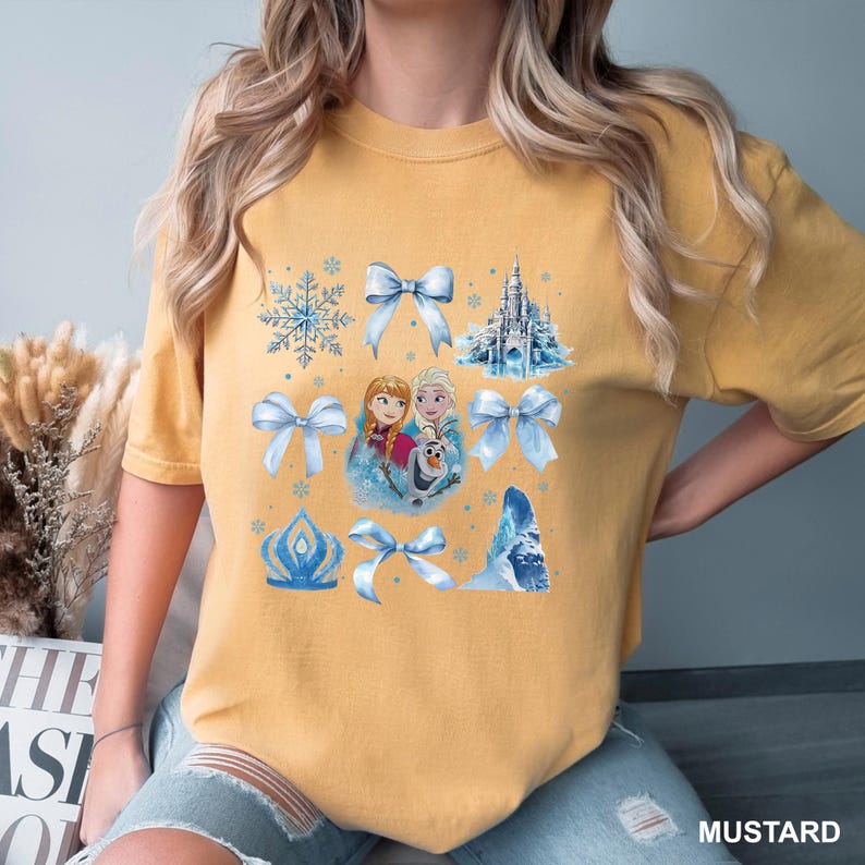 May include: Mustard-colored t-shirt featuring a graphic design with elements from the animated film, including Anna, Elsa, Olaf, a castle, bows, and snowflakes. The word "MUSTARD" is printed at the bottom right.