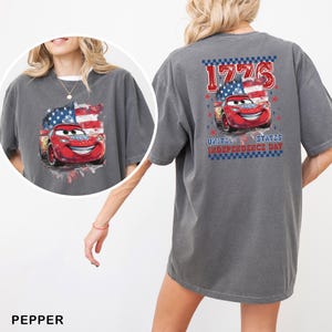 May include: A charcoal grey t-shirt featuring a graphic of a red race car in front of an American flag. The back design includes the text 1776 and United States Independence Day with a chequered border. A circular inset shows the same graphic.