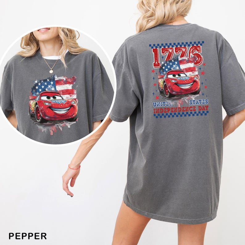 May include: A charcoal grey t-shirt featuring a graphic of a red race car in front of an American flag. The back design includes the text 1776 and United States Independence Day with a chequered border. A circular inset shows the same graphic.