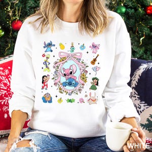 May include: White sweatshirt featuring a colorful graphic of Stitch and other characters. The design includes a pink bow, floral frame, and various cartoon elements. The sweatshirt is a casual style.
